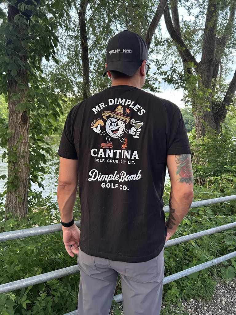 Man wearing Mr. Dimple’s Cantina black t-shirt by Dimple Bomb Golf Co., featuring cartoon golf ball with taco and margarita. Outdoors with trees in background.