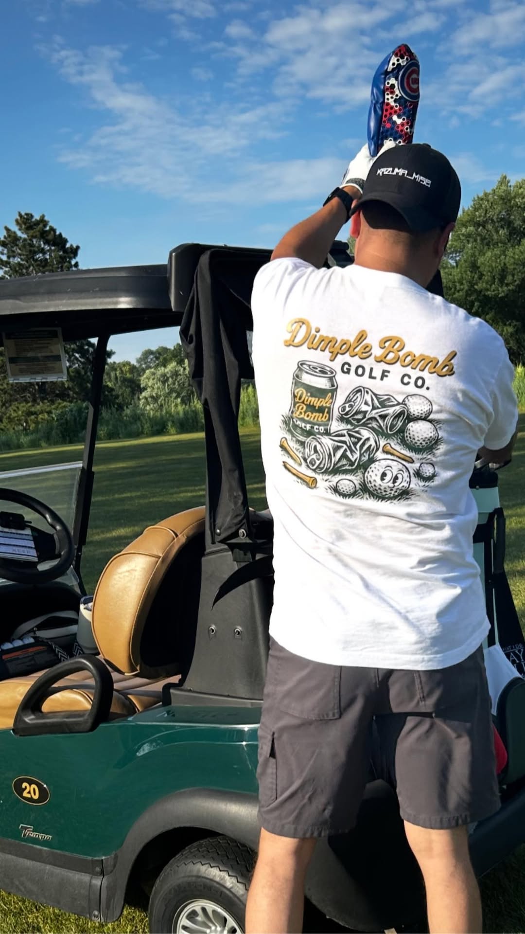 Golfer adjusts club head cover next to a golf cart while wearing the Dimple Bomb 'Crushed Beer' tee, featuring crushed cans and golf balls on the back