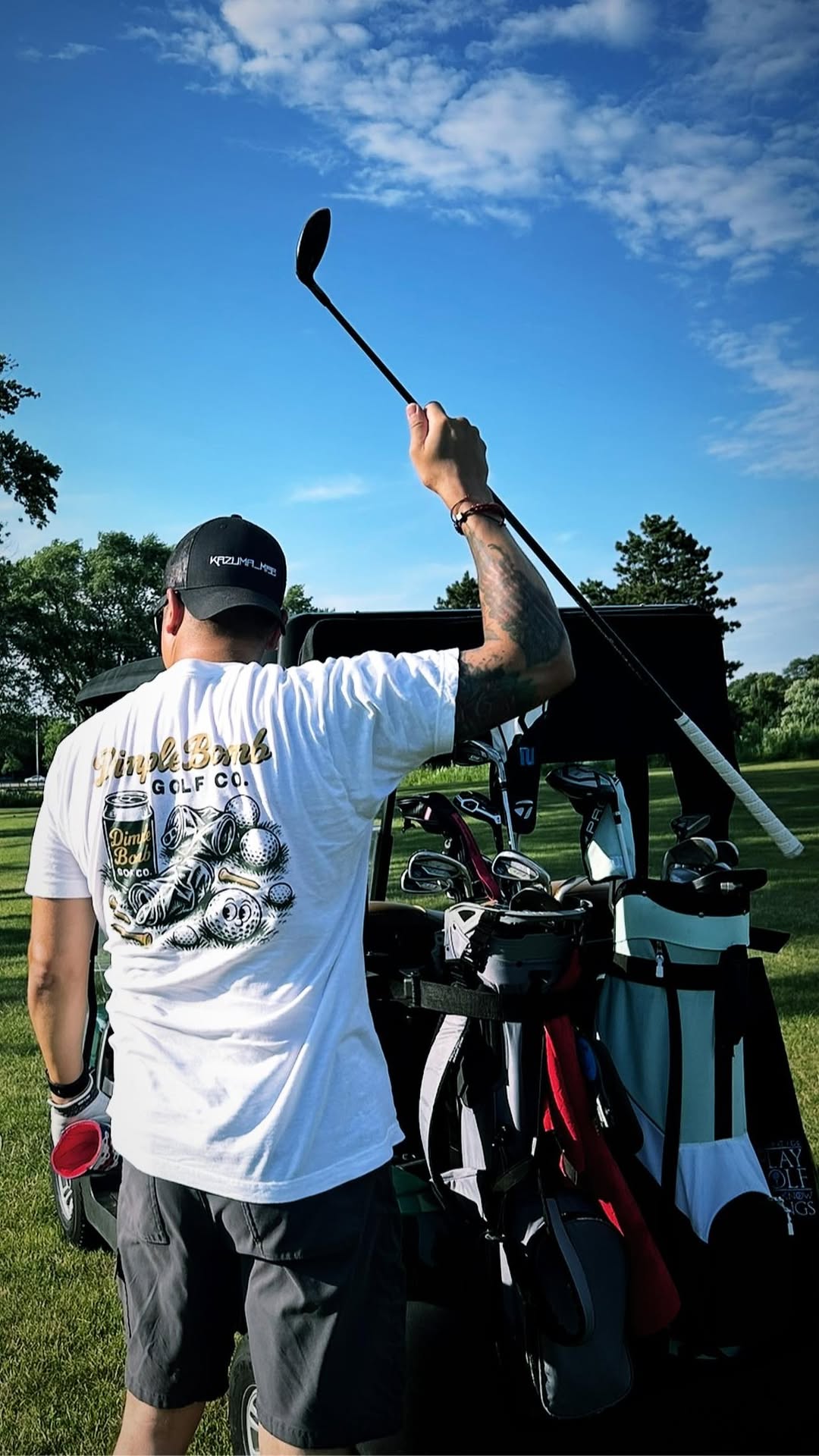 Golfer wearing the Dimple Bomb 'Crushed Beer' tee raises a club triumphantly while standing at the back of a golf cart loaded with gear, under a blue summer sky.