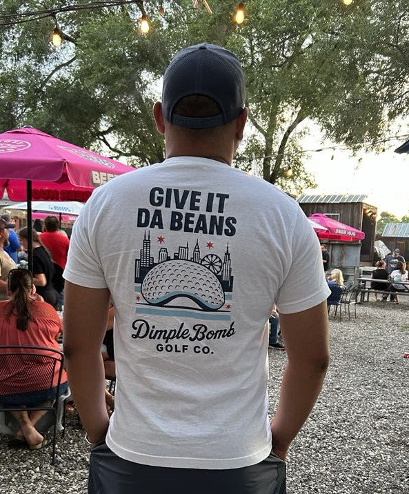 Person wearing a white t-shirt with 'Give It Da Beans' and 'Dimple Bombs Golf Co.' text at an outdoor event.