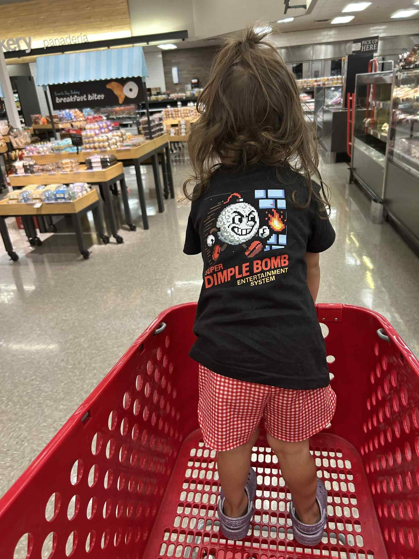 Child in a black Dimple Bomb t-shirt with a 8 Bit pixel graphic design sitting in a red shopping cart inside a store.
