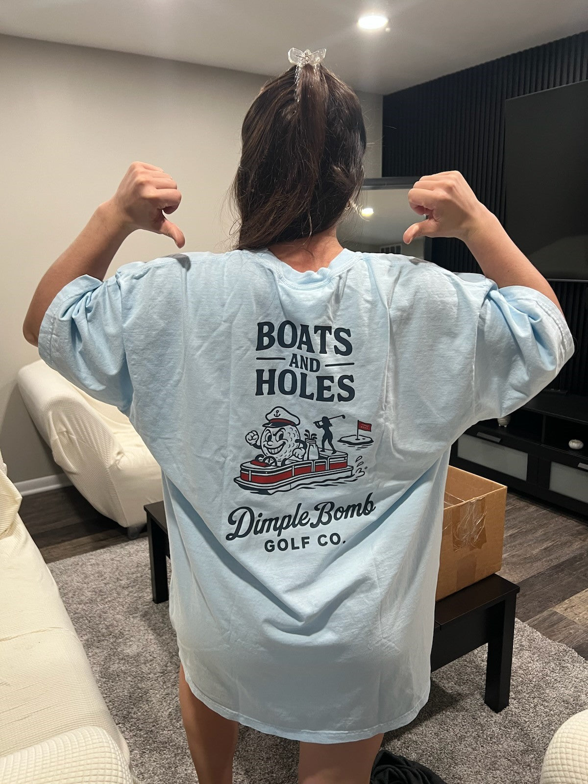 Person wearing a light blue Dimple Bomb Golf t-shirt with 'Boats and Holes' text and graphics in a living room.