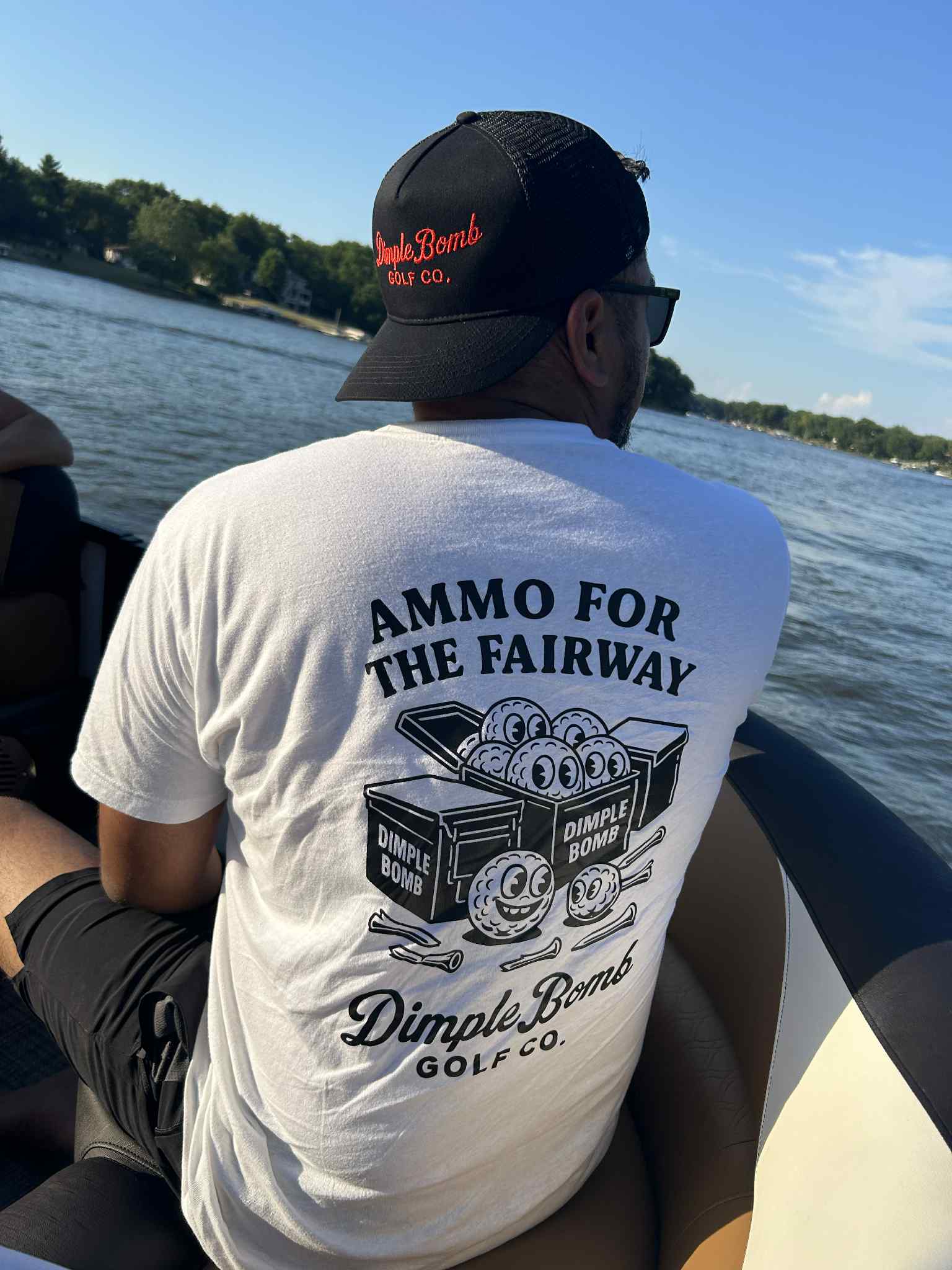 Man wearing "Ammo for the Fairway" golf t-shirt by Dimple Bomb Golf Co. while sitting on a boat, showcasing black ammo can golf ball graphic and mesh trucker hat.
