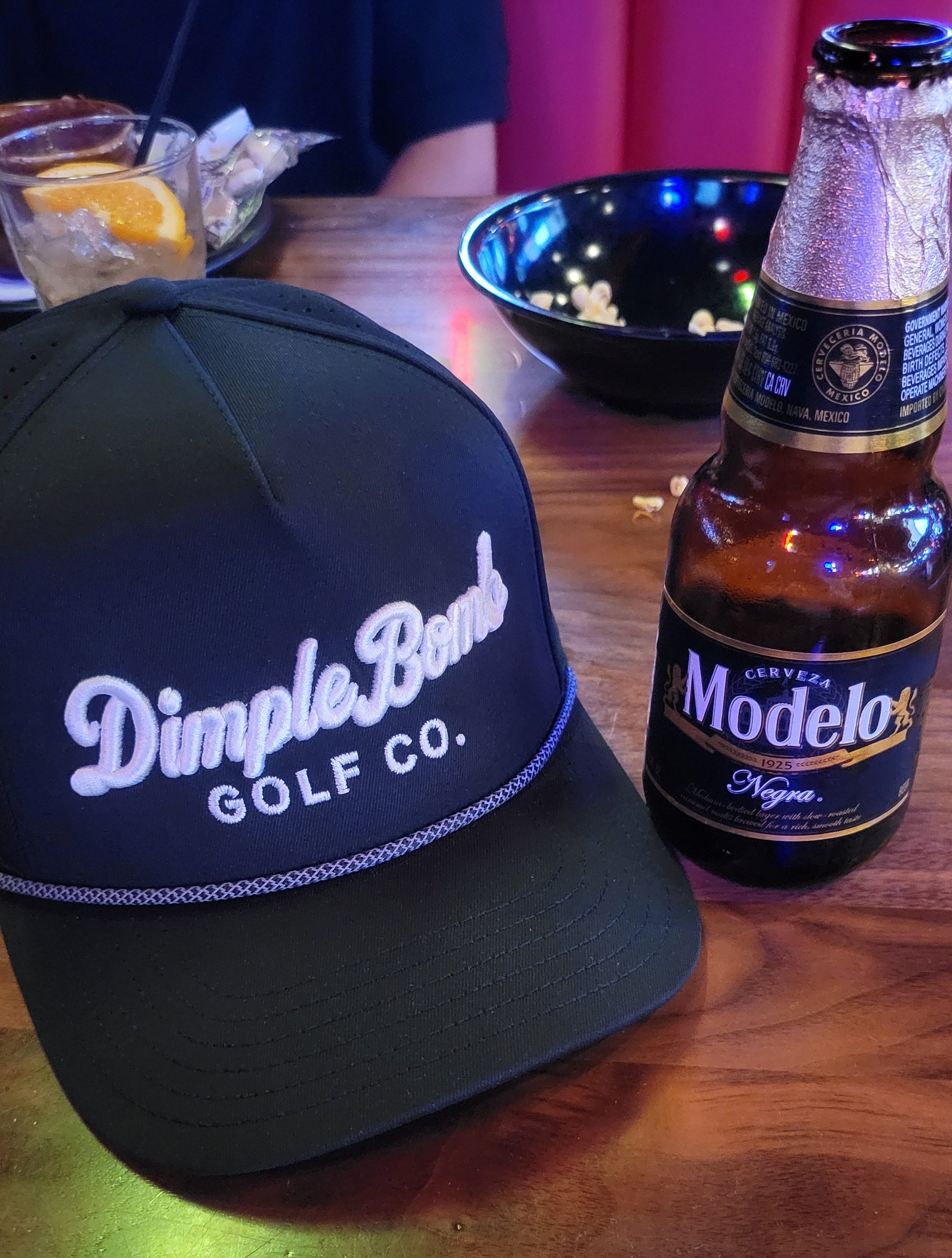 Black golf Hat with 'Dimple Bomb Golf Co' branding next to a Modelo beer bottle on a wooden surface.
