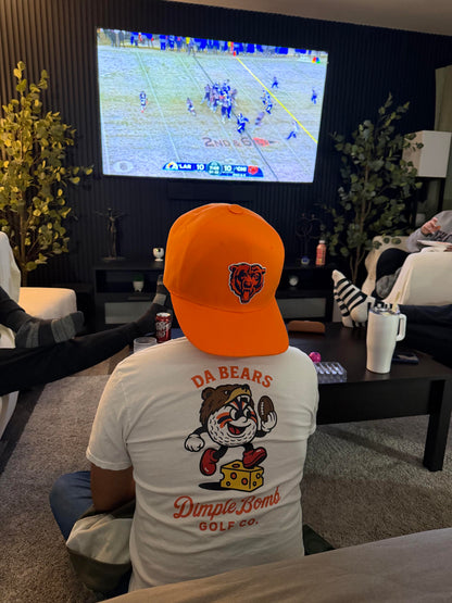 Back view of a white Dimple Bomb Golf Co. Da Bears golf T-shirt featuring Mr. Dimple in a bear hat with a football, worn while watching a Chicago Bears game with an orange Bears hat.