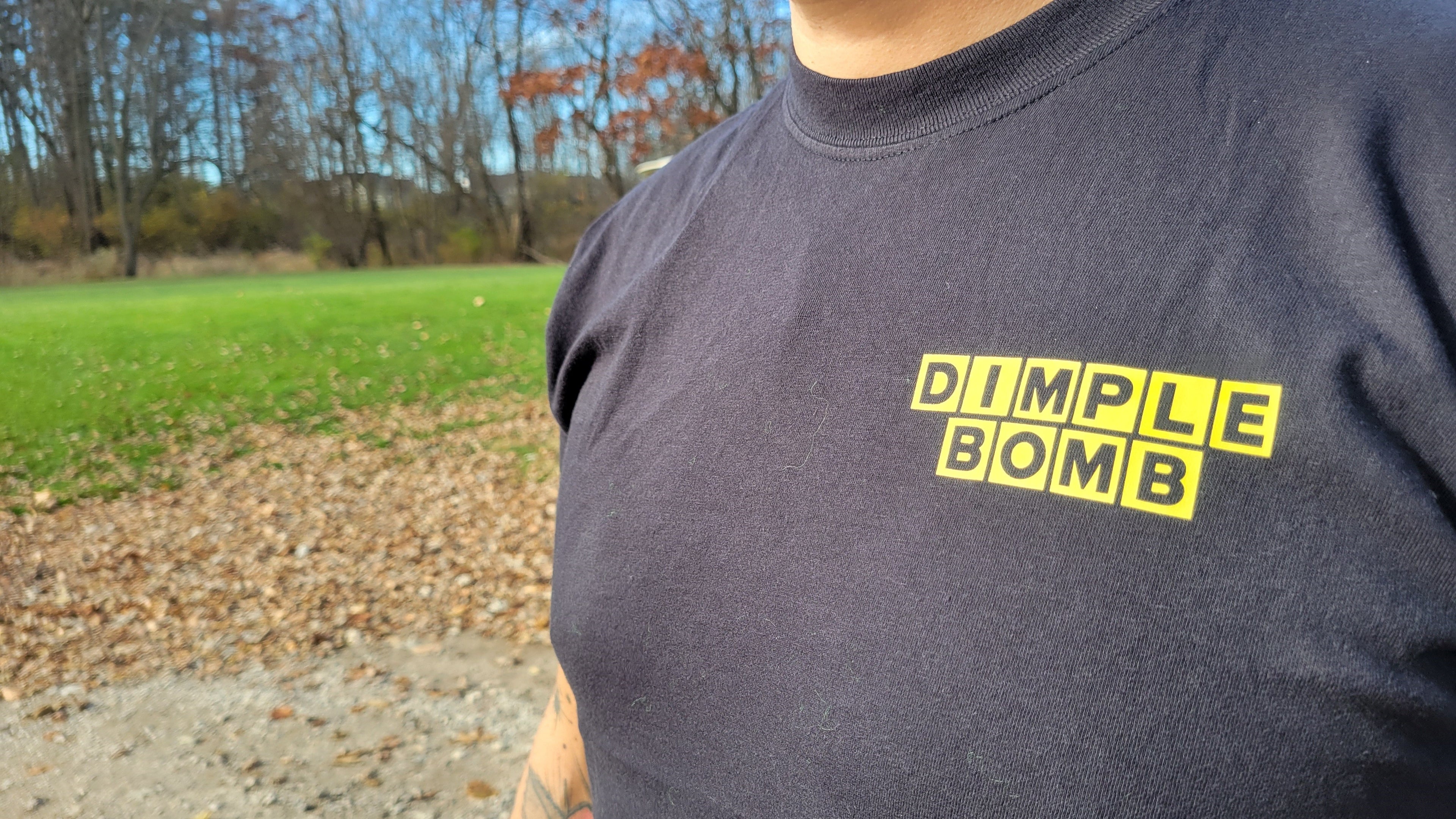 Person wearing a dark t-shirt with 'Dimple Bomb' printed in yellow outdoors.
