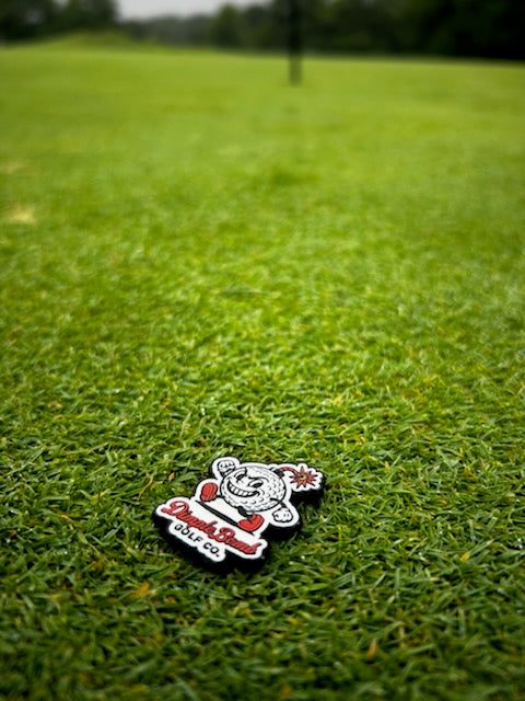 Mr. Dimple golf ball marker featuring the running golf ball character with a lit fuse and Dimple Bomb Golf Co. logo, shown on green grass.
