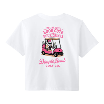 A white short-sleeve t-shirt with a pink graphic on the front. The graphic features a cartoon dog in a pink golf cart with the text 'JUST HERE TO LOOK CUTE AND POUR DRINKS' above it, and 'DimpleBomb GOLF CO.