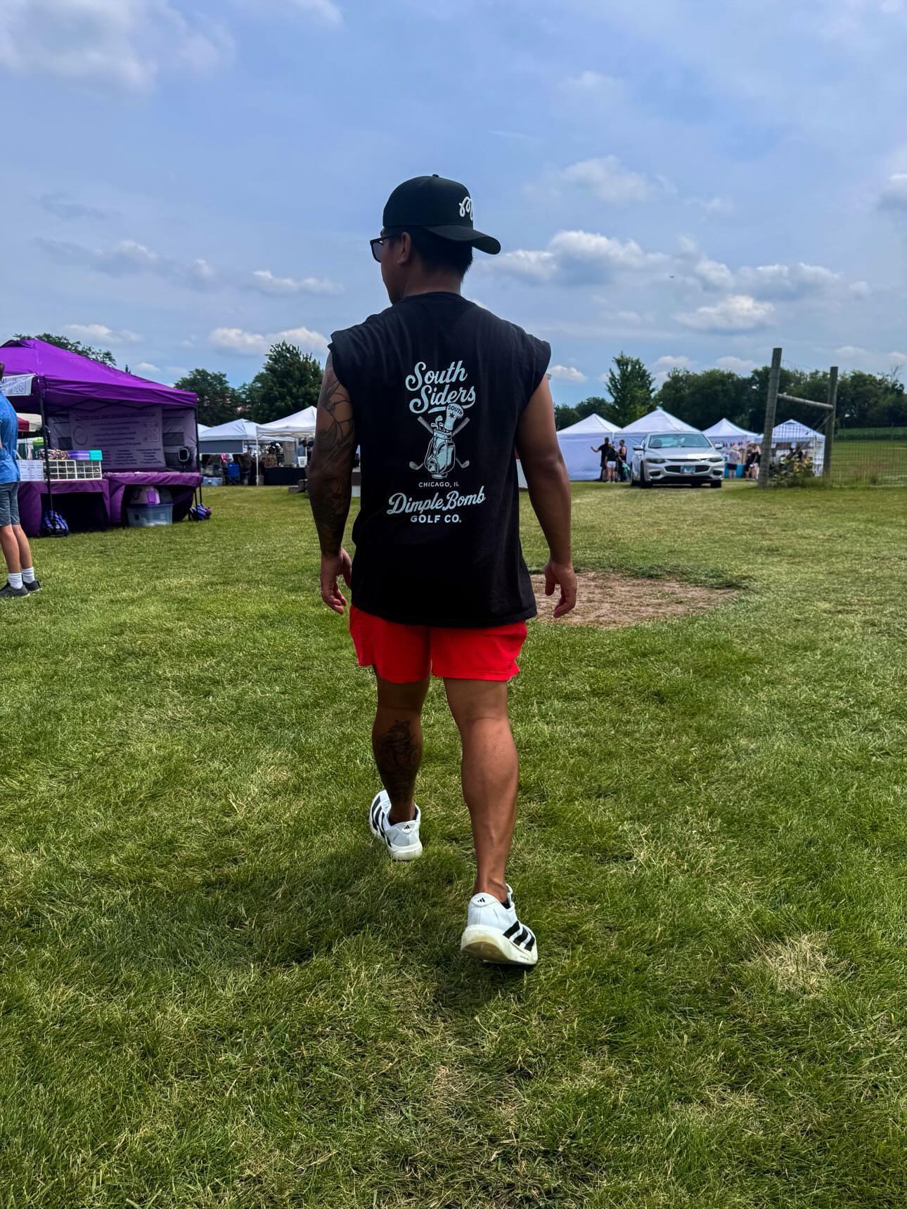 A man walking away on a grassy field at an outdoor festival, wearing a black sleeveless t-shirt with a Black Chicago golf graphic tee featuring a South Siders golf bag design with clubs, a South socks headcover, Chicago, IL text, and the Dimple Bomb Golf Co. logo on the back, red shorts, and white sneakers.